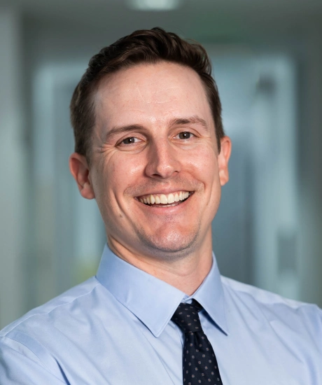 Professional headshot of smiling man in light blue shirt and tie in modern dental office setting.