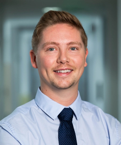 Professional headshot of smiling male dental professional in light blue shirt and dark tie against blurred office background.